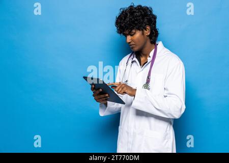 Revolution in healthcare, technologies in medical field. Cheerful positive handsome curly young indian doctor using digital tablet, isolated on blue b Stock Photo