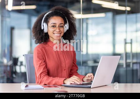 Portrait of young beautiful successful woman at workplace inside office, businesswoman in headphones smiling looking at camera, satisfied with achievement results programmer with laptop. Stock Photo