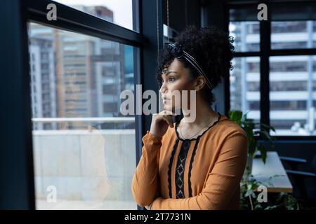 Thoughtful biracial casual businesswoman standing in office looking out of window, copy space Stock Photo