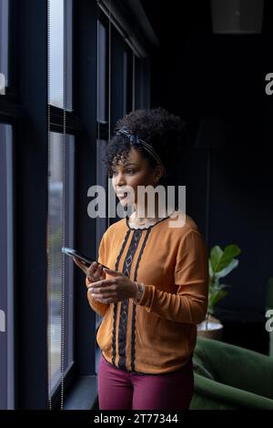 Thoughtful biracial casual businesswoman using smartphone, looking out of office window, copy space Stock Photo