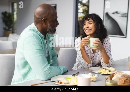 Happy mature diverse couple having breakfast and coffee in sunny kitchen Stock Photo