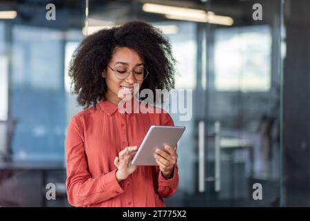 Young successful smiling female programmer inside office at workplace, satisfied woman holding tablet computer in hands, testing new software, business woman with curly hair and shirt. Stock Photo