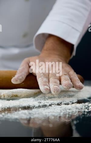 Close up of a chef hand covered in flour rolling a rolling pin across a dough over a glass table Stock Photo