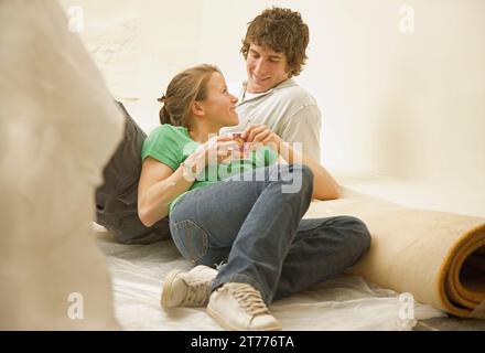 Couple having a break from DIY lying on the floor leaning on a rolled up carpet and looking at each Stock Photo