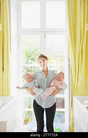 Smiling Mother Holding Twin Babies in Nursery Stock Photo