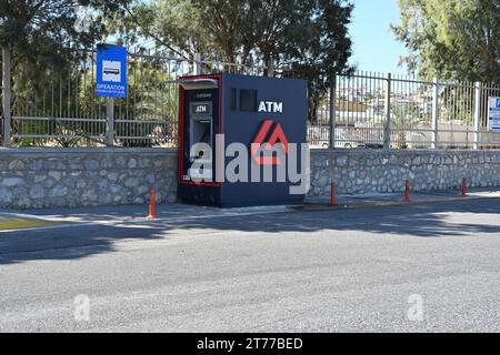 Blue ATM machine of Eurobank is standing near bus stop sign in the area of passenger seaport. Behind is metal fence and small stone wall. Stock Photo