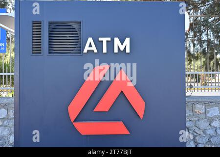 Side view on blue ATM machine of Eurobank standing alone in the area of passenger seaport. Behind is metal fence and small stone wall. Stock Photo