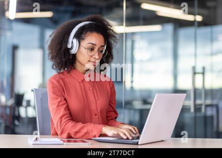 Serious concentrated woman working with laptop inside office, businesswoman thinking with headphones typing on keyboard, programmer developer listening to audio podcast, training course. Stock Photo