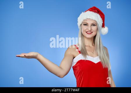 Young Santa woman in red costume and cap presenting an invisible product to the copyspace, isolated on blue background Stock Photo