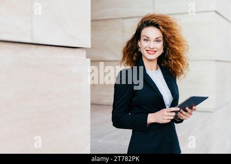 Professional woman with curly hair, smiling, holding a tablet, stands against a cream-colored wall, exuding confidence and approachability Stock Photo