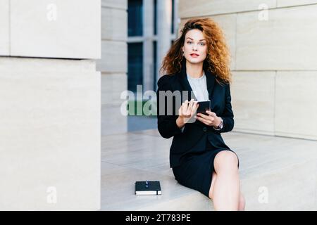 Contemplative businesswoman with striking curly hair using a smartphone, seated by a modern building, thoughtful Stock Photo