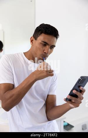 Biracial man brushing teeth and using smartphone in bathroom at home Stock Photo