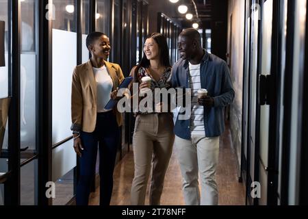 Happy diverse male and female colleagues with coffees, tablet and smartphone talking in corridor Stock Photo