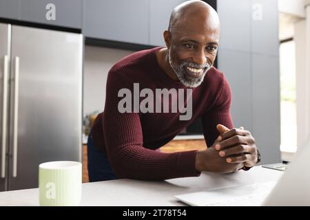Happy mature african american man having coffee using laptop standing in sunny kitchen Stock Photo