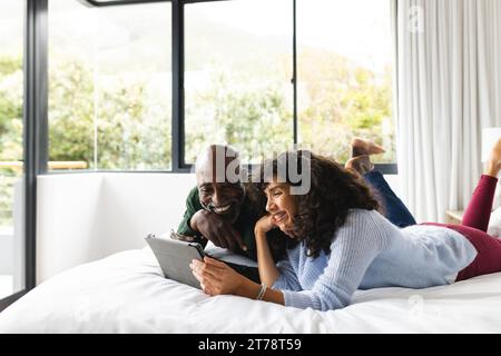 Happy diverse mature couple lying on bed using tablet in sunny bedroom, copy space Stock Photo