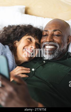 Happy diverse mature couple lying on bed embracing using tablet in sunny bedroom Stock Photo