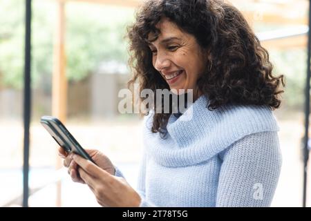 Happy mature caucasian woman using smartphone smiling by window at home Stock Photo