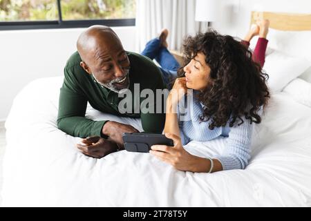 Happy diverse mature couple lying on bed using smartphone, talking in sunny bedroom Stock Photo