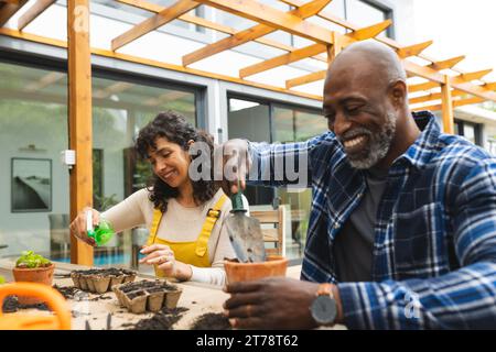 Happy mature diverse couple potting and watering seedling plants on garden terrace Stock Photo