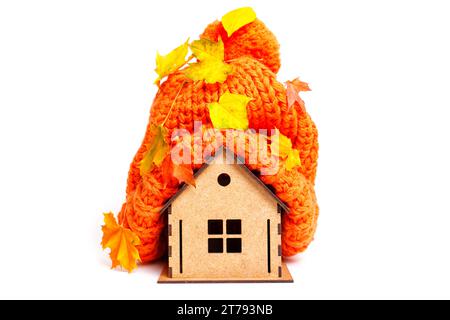 Miniature house model with a thick orange hat placed on top and covered with pigmented leaves is isolated on white background. Getting ready for a col Stock Photo