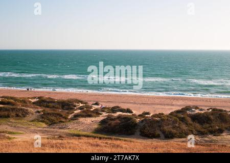 Seascape. Blue sea and sandy shore with dunes on an autumn day. Tourists walking in the background. Stock Photo