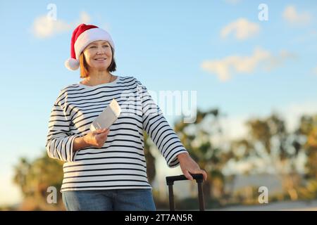 Independent mature woman in Christmas Santa Claus hat, striped shirt holds passport, boarding ticket, suitcase on seashore with palms backdrop celebra Stock Photo