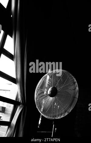 Black and white photo of a vintage floor fan standing by a tall window in a loft. Stock Photo