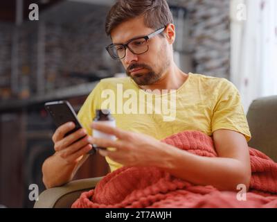 Young Caucasian man using smart phone for reading, searching prescription on bottle medicine, pill label text about information online, instructions side effects, pharmacy medicament concept Stock Photo