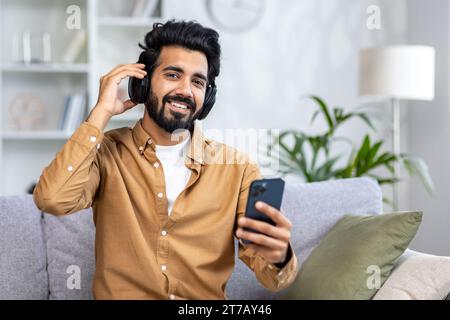 Satisfied Arab man with a beard in headphones sitting on the sofa listening to music, holding a phone in his hand, taking a selfie, dancing to his favorite music, looking at the camera. Stock Photo