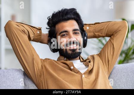 Portrait of happy hindu man wearing headphones relaxing on sofa in living room at home. Smiling student listens to his favorite podcast music, puts hands behind his head, smiles and looks at camera. Stock Photo