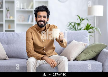 Happy sincere Arab man sitting on sofa at home holding cup in hands, drinking tea or water, concept of healthy lifestyle, healthy habits, rest after working day, smiling looking at camera. Stock Photo