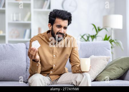 Sick Hindu man sitting on sofa at home, holding cup in hand, drinking tea, holding napkin, viral diseases, runny nose and cough, health concept. Stock Photo