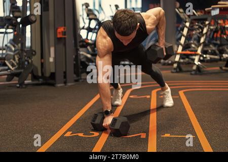 Athlete planking with dumbbells in fitness gym. Athletic man doing gym dumbbell plank row. Fitness workout. Developing fitness and core strength. Stock Photo