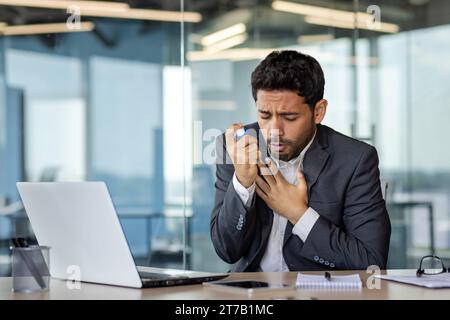 Arab businessman suffering from asthma in modern office, young man holding inhaler in hand and holding chest, breathing hard, breathing and health problems Stock Photo