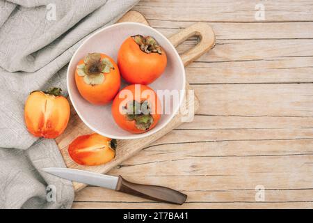 Fresh ripe persimmons in white bowl on cutting board on wooden table. Healthy vegan food concept. Top view, flat lay, mockup. Stock Photo