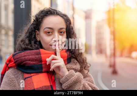 ethnic curly-haired woman in a jacket and an orange jacket holding her index finger to her lips. Shushing concept Stock Photo