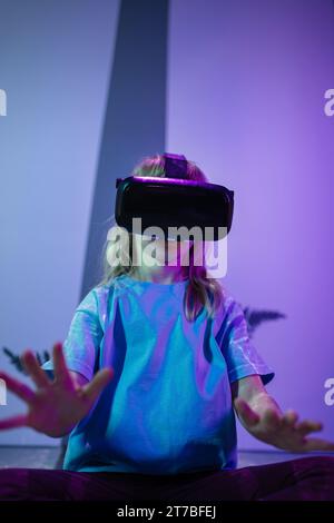 School aged girl child in VR glasses sitting on the floor, having an immersive experience through interaction with virtual reality. Stock Photo