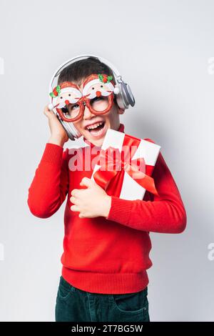 Happy boy Generation Alpha in a red christmas holidays sweater and funny party glasses listen to music on headphones. Stylish boy Gen Alpha with a Stock Photo