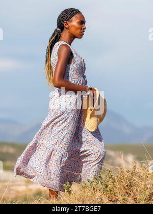 Full body portrait of an expressive young African woman in a dreamy dress holding a hat and looking at the Atlantic Ocean. Stock Photo
