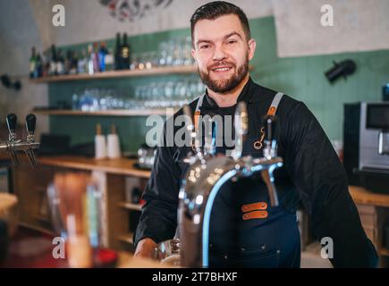 Portrait of happy smiling bearded barman dressed in a black uniform with an apron at bar counter with draught beer taps. Successful people, hard work, Stock Photo
