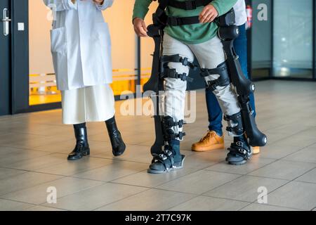 Mechanical exoskeleton, female doctor with unrecognizable engineer walking with disabled person with robotic skeleton in rehabilitation Stock Photo