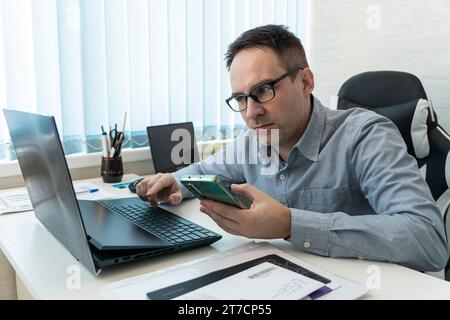 Handsome young businessman working with laptop computer at his desk in office. Happy male entrepreneur typing on pc keyboard, checking email, taking p Stock Photo