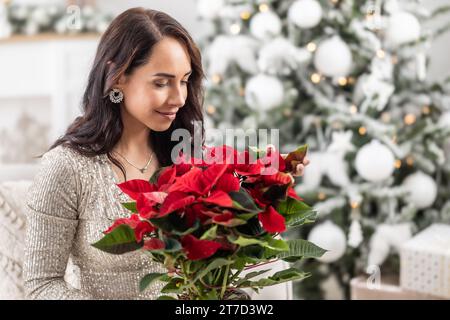 Woman smiles and smells a poinsette in a flower pot next to a Christmas tree. Stock Photo