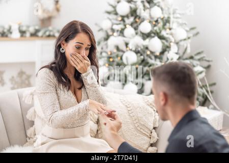 Happily crying woman lets her proposing boyfriend put a ring on her finger during Christmas. Stock Photo