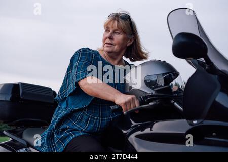 Female biker. Independent mature blonde is having great time, looking away while sitting on her black motorcycle on autumn day in nature. Hobby - moto Stock Photo