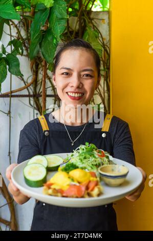 Young vietnamese waitress holding a plate with eggs bennedict waffles with smoked salmon in restaurant Stock Photo