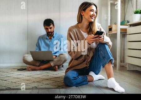 Smiling woman using smart phone with man sitting with laptop. Social media home office concept. Stock Photo