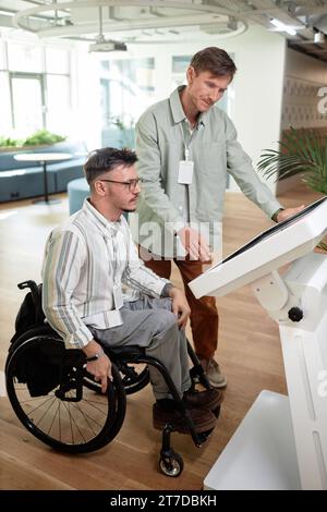 Vertical image of man helping to make online entry for man with disability in office hall Stock Photo