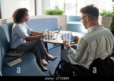 Rear view of young businessman with disability working on tablet pc while sitting at table with his colleague Stock Photo
