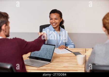 Smiling young businesswoman shaking hand with her colleague while they sitting at table at meeting Stock Photo